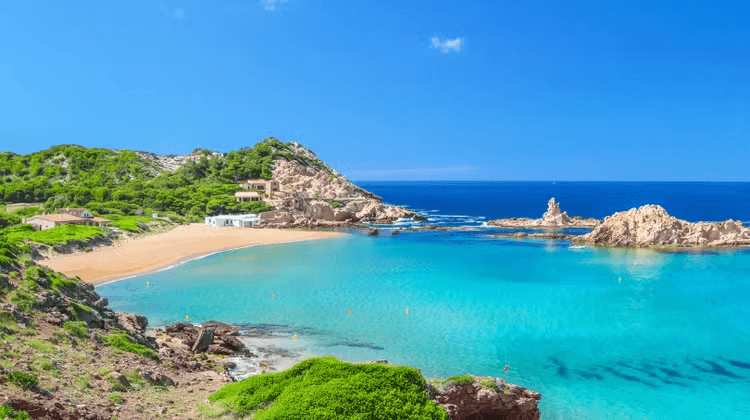 türkisfarbene Badebucht der Balearen in Menorca vor einer felsigen Küstenlandschaft mit kristallklarem Mittelmeer. Die geschützte Ankerbucht bietet ideale Bedingungen für Yachtcharter auf Menorca und einen exklusiven Segelurlaub entlang der malerischen Inselküsten der Balearen unter strahlend blauem Himmel.