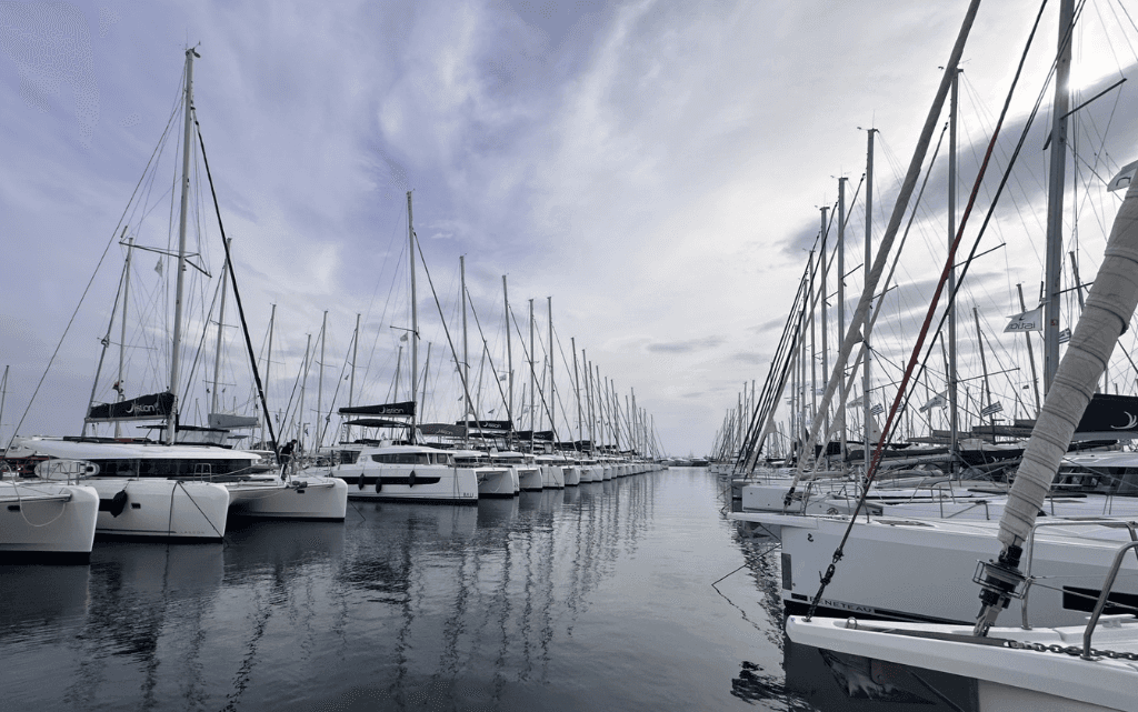 Eine ruhige Marina erstreckt sich unter einem leicht bewölkten Himmel, in der zahlreiche Segelboote dicht nebeneinander an beiden Seiten eines schmalen Wasserkanals festgemacht sind. Die schlanken Masten der Boote ragen in den Himmel und bilden ein dichtes, fast geometrisches Muster aus Linien. Das Wasser ist glatt und spiegelt die Boote sowie die Masten leicht verschwommen wider. Die Szene wirkt still und geordnet, mit einer fast symmetrischen Perspektive, die den Blick in die Tiefe des Hafens lenkt. Es herrscht eine entspannte, beinahe meditative Atmosphäre ohne sichtbare Bewegung.