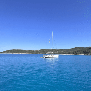 Ein einzelnes Segelboot liegt ruhig auf einem türkisblauen, klaren Meer. Das Wasser ist so still, dass sich das Boot leicht darin spiegelt. Im Hintergrund erstreckt sich eine grüne, hügelige Küstenlandschaft unter einem wolkenlosen, strahlend blauen Himmel. Die Atmosphäre wirkt friedlich und beinahe zeitlos, als wäre das Boot fernab von jeglicher Hektik unterwegs.