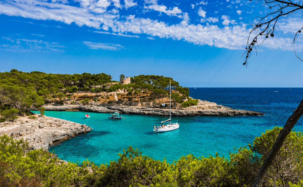 Weiße Segelyacht in einer türkisfarbenen Badebucht der Balearen vor einer felsigen Küstenlandschaft mit Pinienwäldern und kristallklarem Mittelmeer. Die geschützte Ankerbucht bietet ideale Bedingungen für Yachtcharter auf Mallorca und einen exklusiven Segelurlaub entlang der malerischen Inselküsten der Balearen unter strahlend blauem Himmel.