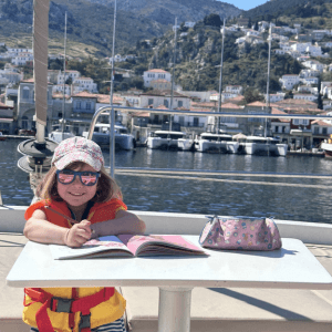 Ein Kind sitzt an einem kleinen Tisch an Bord eines Bootes und malt oder schreibt konzentriert in ein aufgeschlagenes Heft. Es trägt eine orangefarbene Schwimmweste sowie eine helle Kappe mit Muster und eine Sonnenbrille mit getönten Gläsern, die das Gesicht vor der Sonne schützt. Das Kind lächelt leicht und wirkt entspannt und zufrieden bei seiner Beschäftigung. Auf dem Tisch liegt neben dem Heft ein Federmäppchen mit buntem Design. Im Hintergrund erstreckt sich ein malerischer Hafen mit mehreren festgemachten Booten und Yachten, dahinter steigen dicht bebaute Häuser an einem grünen Hügel hinauf. Die Szene vermittelt eine Mischung aus Urlaubsstimmung, Kreativität und entspanntem Leben am Wasser.