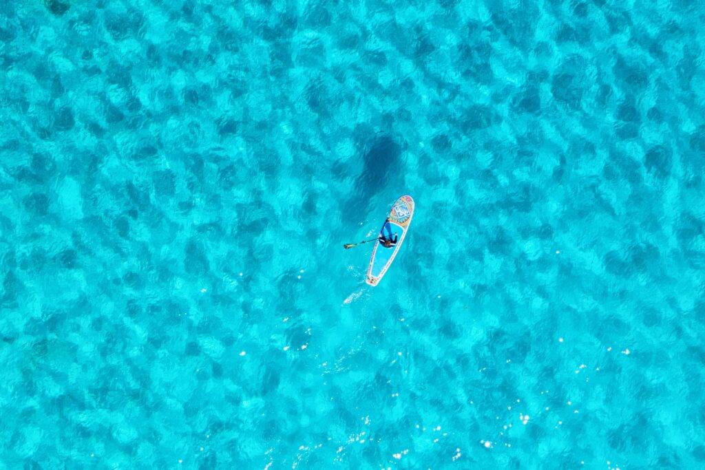 Stand-up-Paddler auf kristallklarem türkisfarbenem Wasser der Balearen, von oben fotografiert – mediterranes Urlaubsparadies für Segelreisen.