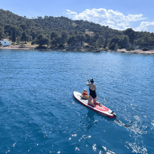 Eine Person steht auf einem Stand-up-Paddle-Board und paddelt über das klare, tiefblaue Wasser. Vor ihr sitzt ein Kind, beide tragen Schwimmwesten. Die Umgebung zeigt eine ruhige Bucht mit bewaldeten Hügeln und wenigen Gebäuden entlang der Küste. Das Wasser glitzert im Sonnenlicht, und die Szene vermittelt ein Gefühl von Freizeit, Abenteuer und gemeinsamer Aktivität in der Natur.