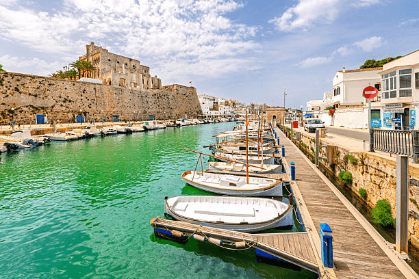Charmanter Hafen der Balearen mit traditionellen Booten entlang einer historischen Uferpromenade und türkisgrünem Wasser. Die maritime Kulisse eignet sich perfekt für Yachtcharter, Segelyachten und Segelreisen rund um Mallorca, Menorca und die weiteren Baleareninseln.