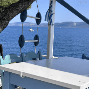 Ein weiß gestrichener Tisch und eine Bank stehen auf einer Terrasse mit Blick auf das Meer. Daneben hängt eine dekorative Leiter mit runden Elementen, und eine griechische Flagge weht sanft im Wind. Im Hintergrund ist das offene Meer zu sehen, auf dem ein Segelboot vorbeifährt, sowie entfernte Inseln oder Küstenlinien. Die Szene strahlt Ruhe, Einfachheit und mediterranes Flair aus.