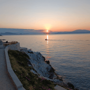 Die Sonne geht langsam über dem Meer unter und taucht den Himmel in warme Orangetöne. Ein kleines Boot fährt in der Ferne über das ruhige Wasser und hinterlässt eine feine Spur. Im Vordergrund verläuft ein schmaler Weg entlang einer felsigen Küste mit niedriger Vegetation und vereinzelten Blumen. Die Silhouetten von Bergen oder Inseln sind am Horizont zu erkennen und verstärken die ruhige, romantische Stimmung des Moments.