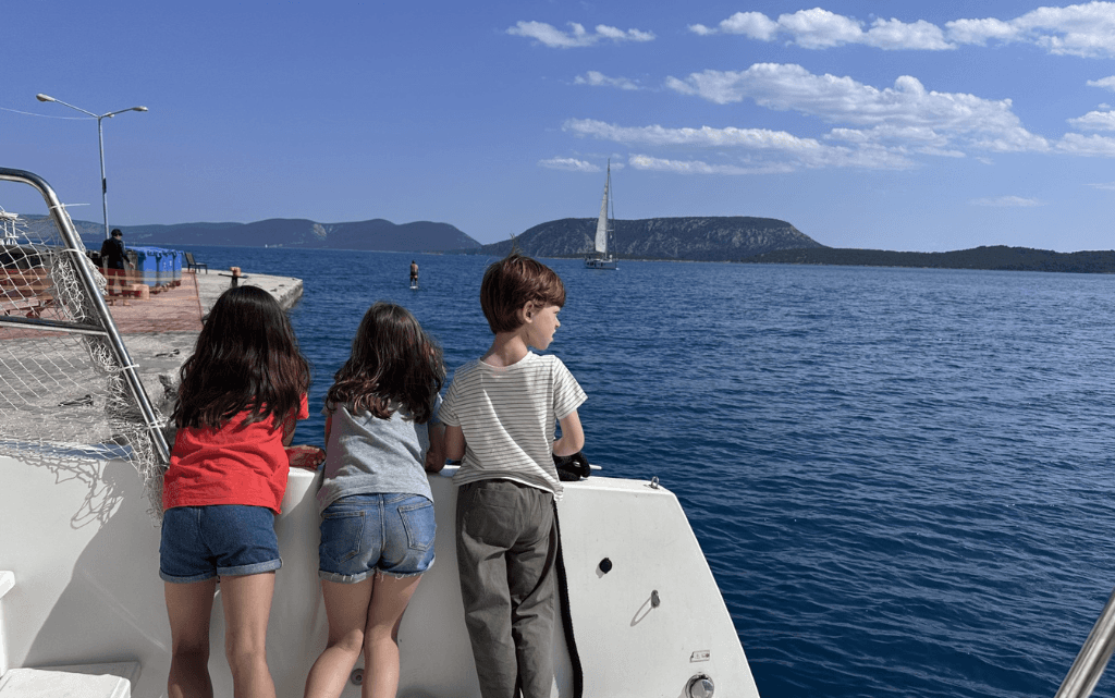 Drei Kinder lehnen sich über die Reling eines Bootes und blicken neugierig auf das Wasser hinaus. Sie tragen sommerliche Kleidung wie T-Shirts und Shorts. Das Meer ist tiefblau und ruhig, während im Hintergrund ein Segelboot vorbeifährt und sich eine hügelige Küstenlandschaft erstreckt. Die Körperhaltung der Kinder zeigt Interesse und Begeisterung, als würden sie etwas im Wasser beobachten oder einfach die Aussicht genießen.